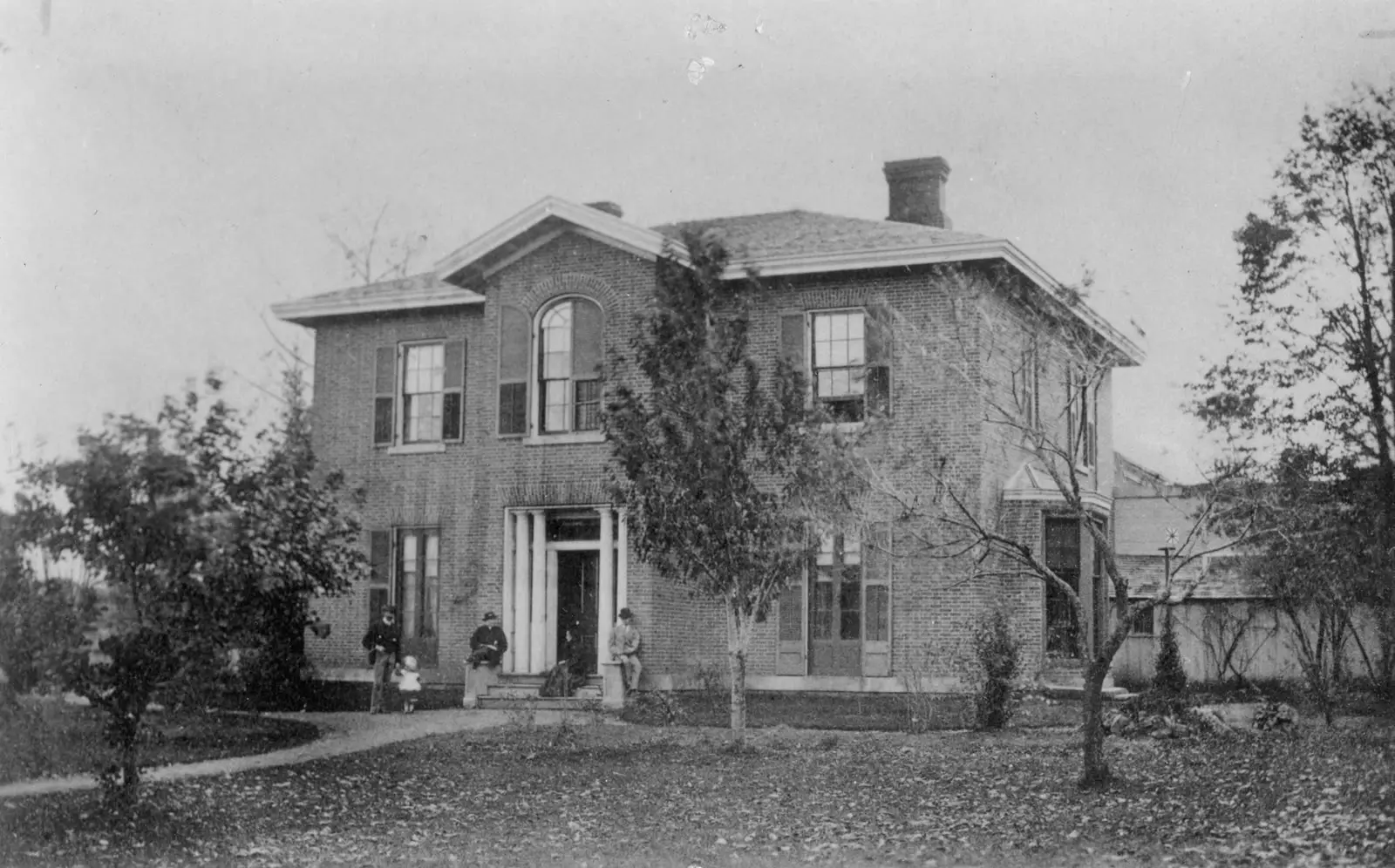 A two-storey brick house with a central gable, an arched upper window, and a columned doorway, with a family standing on the front walk
