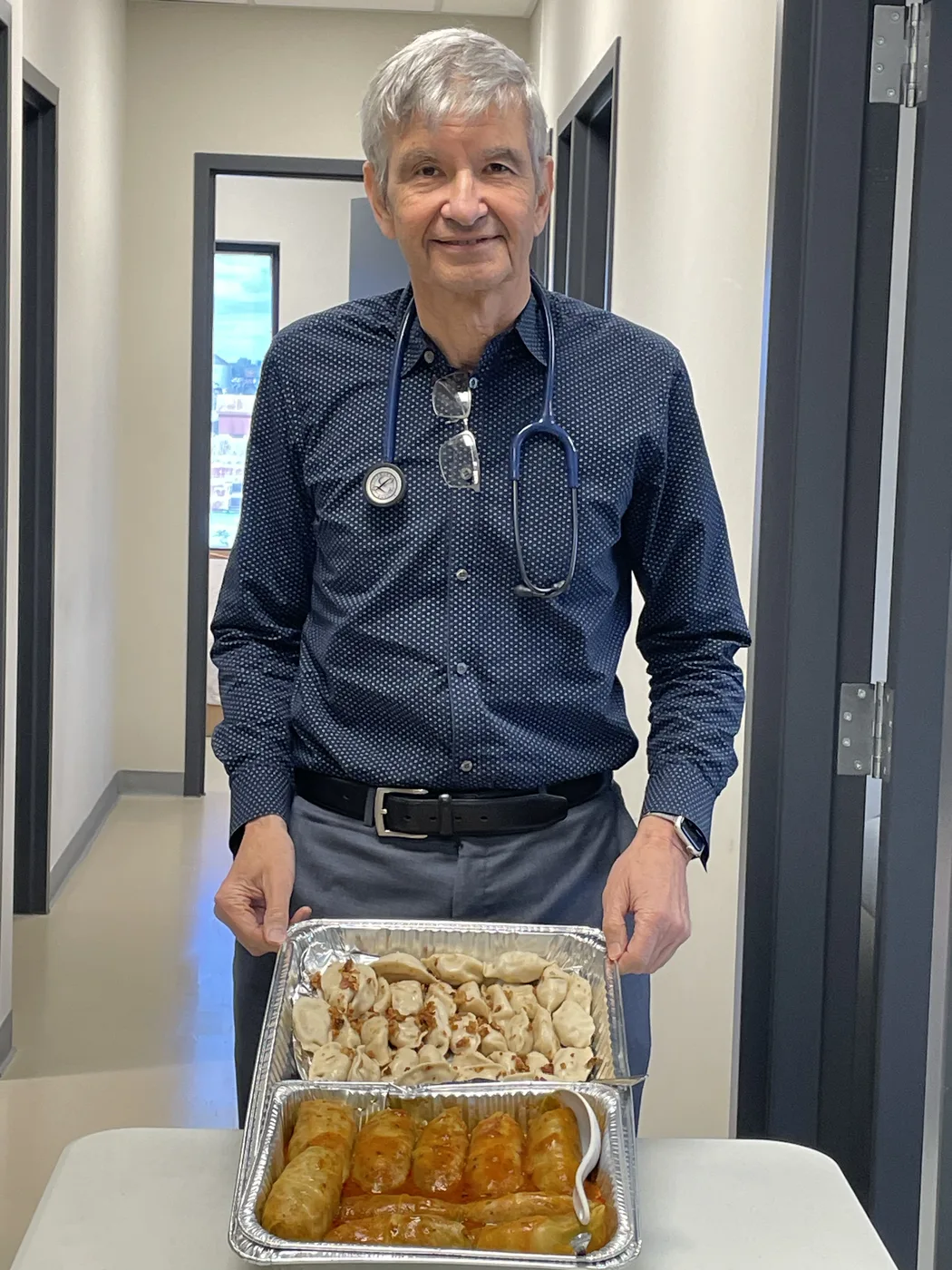 Dr. Rick Tytus in a clinic hallway, wearing a dress shirt and stethoscope, carrying two large foil catering trays of homemade Polish food